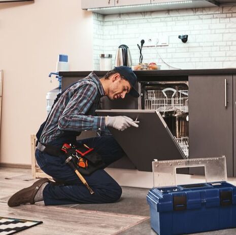 Technician wearing tool belt sitting on kitchen floor working dishwasher repairs. Blue toolbox beside man.